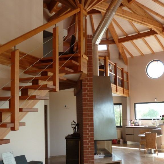 Escalier en bois dans une maison moderne avec poutres apparentes, cheminée en briques et fenêtre ronde.