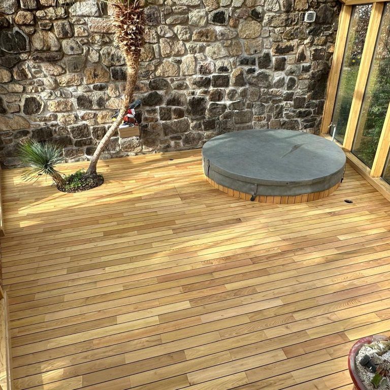 Terrasse en bois avec jacuzzi, palmier et mur en pierre.