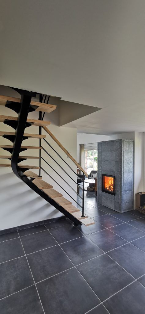 Point de vue éloigné sur le poêle à bois en pierre ollaire Mammutti de Tulikivi de forme rectangulaire au milieu d'un salon avec foyer allumé et escalier devant