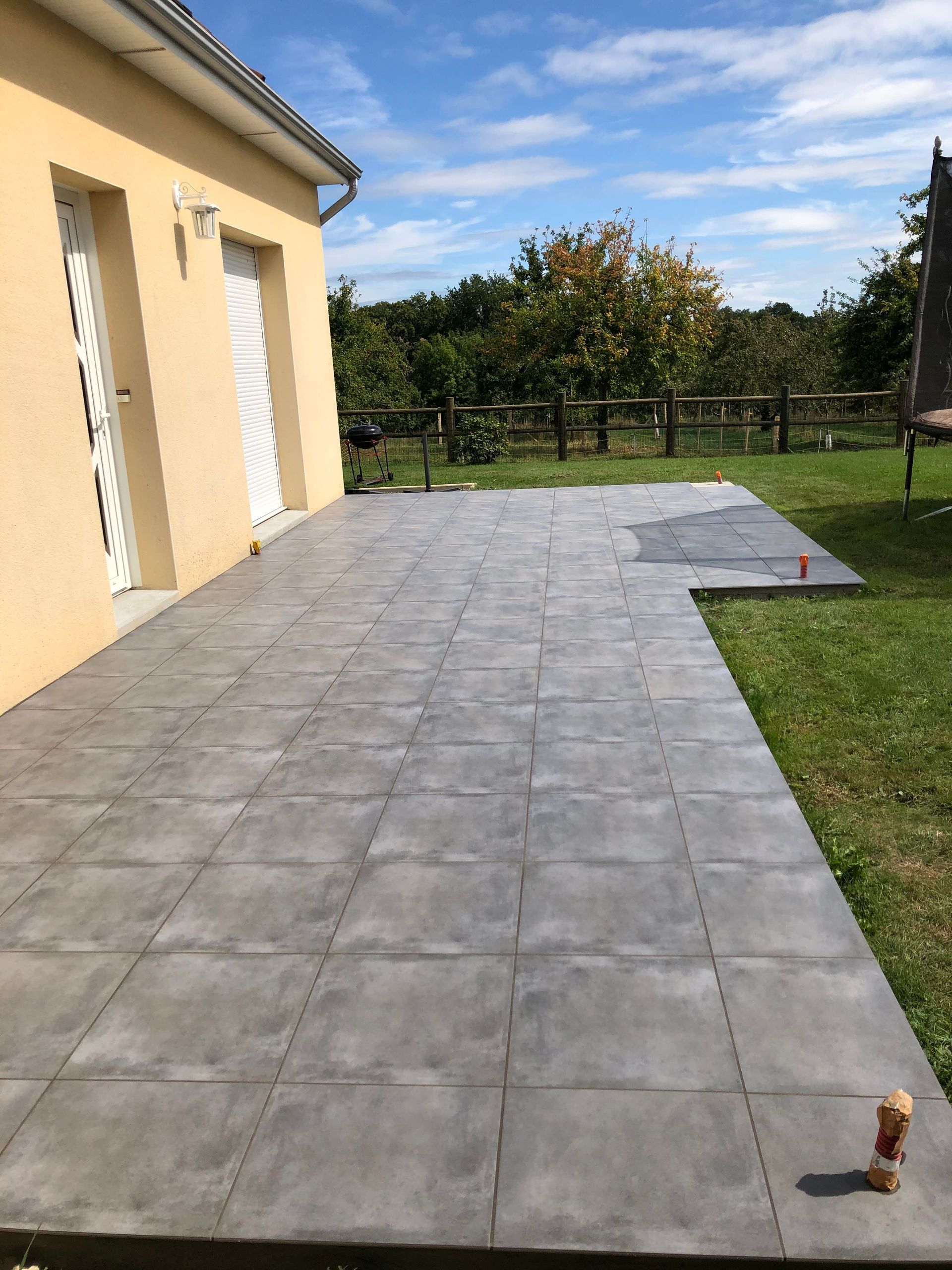 Terrasse carrelée extérieure jouxtant une maison beige, bordée de pelouse et d'une clôture sous un ciel partiellement nuageux.
