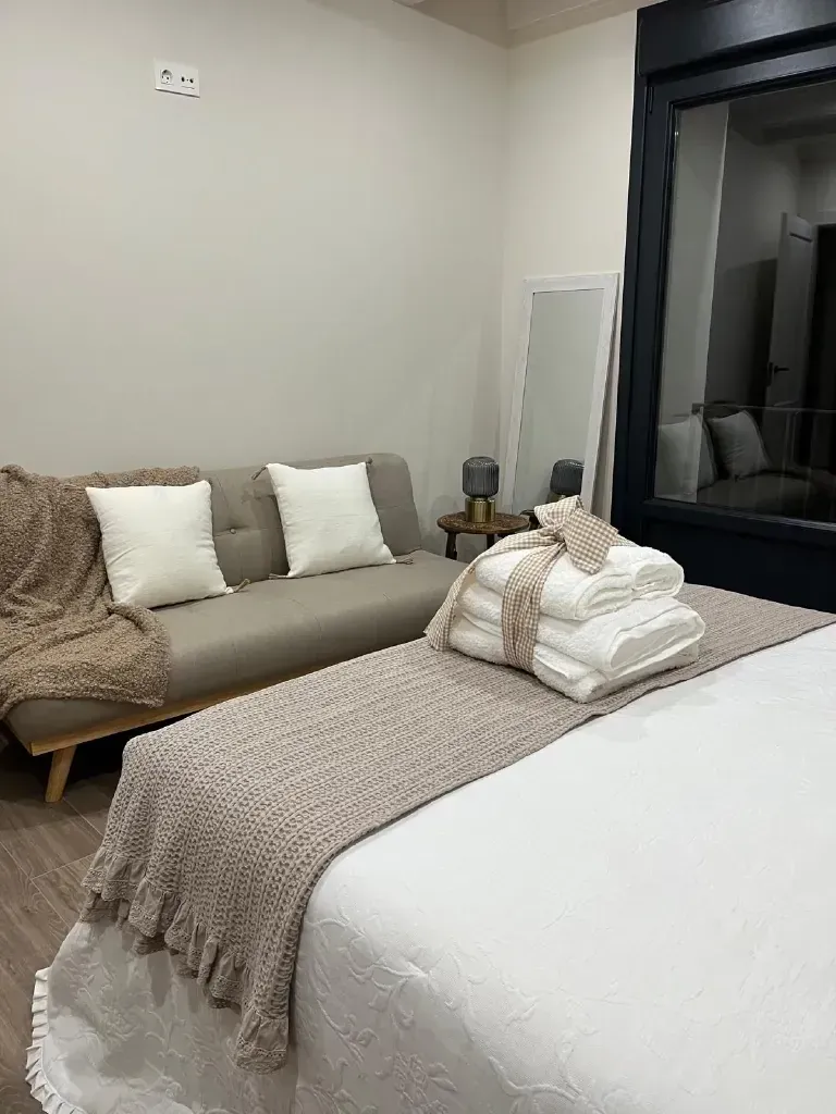 Dormitorio con cama, sofá, toallas y espejo. Predominan los tonos beige, blanco y marrón.