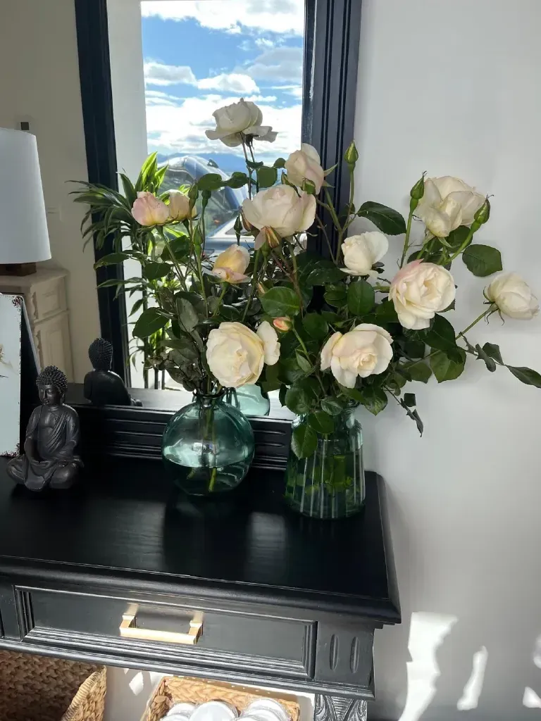 Rosas blancas en jarrones de cristal sobre una mesa negra con un espejo que refleja una vista panorámica.
