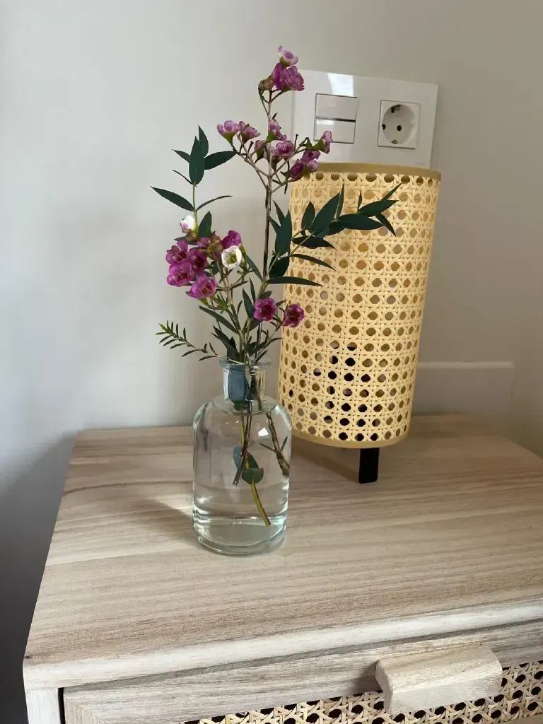 Un jarrón con flores rosas y blancas sobre una mesita de noche de madera junto a una lámpara tejida.
