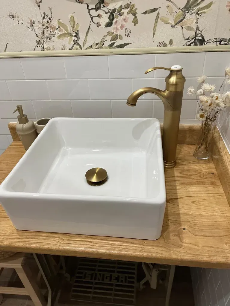 Lavabo rectangular blanco con grifo dorado sobre encimera de madera, fondo de papel tapiz floral.