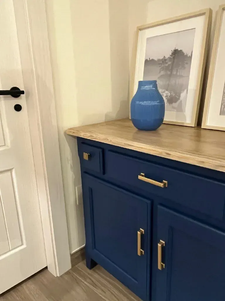 Gabinete azul con herrajes dorados, tapa de madera clara, jarrón azul, fotografías enmarcadas y una puerta blanca.