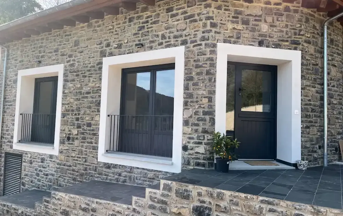 Exterior de casa de piedra con puertas y marcos de ventanas oscuros, molduras blancas y un pequeño escalón hasta la entrada.