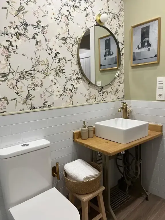 Baño con papel pintado floral, espejo redondo, lavabo blanco sobre superficie de madera e inodoro.