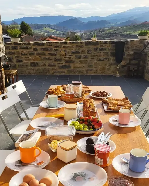 Mesa de desayuno en un patio con vistas a la montaña. Incluye bollería, fruta y huevos.