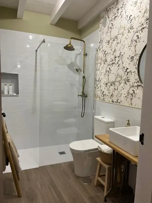 Baño con ducha de azulejos blancos, papel tapiz floral, inodoro, lavabo y piso de madera.