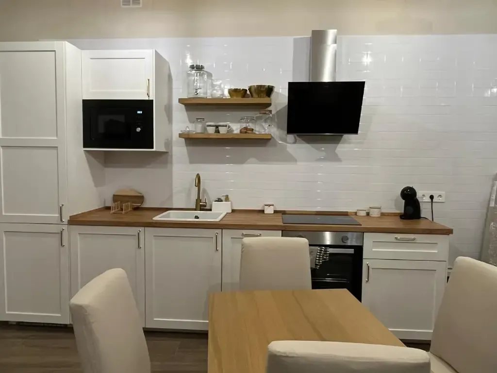 Cocina blanca con encimeras de madera, estantes y mesa; microondas y horno visibles.