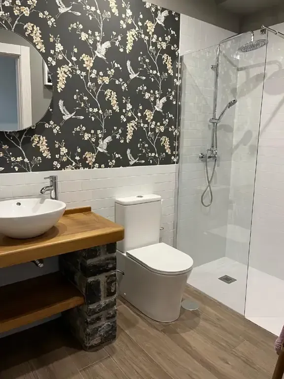 Baño con papel pintado floral, ducha de cristal, tocador de madera e inodoro blanco.