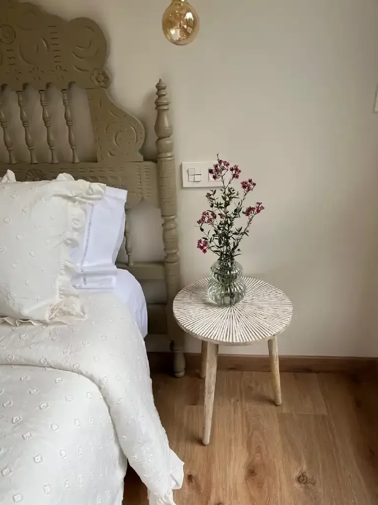 Dormitorio con cabecero tallado, ropa de cama blanca y una mesa auxiliar con flores.