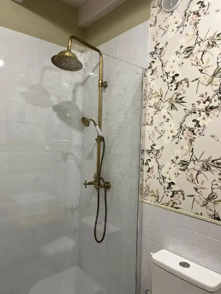 Ducha con accesorios de latón antiguo, azulejos blancos, papel tapiz floral y un inodoro.