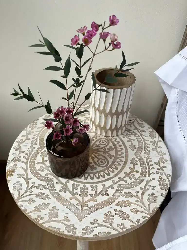Pequeña mesa redonda con diseño tallado, dos jarrones con flores rosas y ropa de cama blanca.