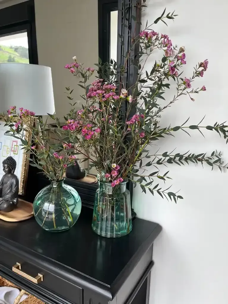 Dos jarrones de cristal con flores rosas sobre una mesa negra, con una lámpara y un espejo al fondo.