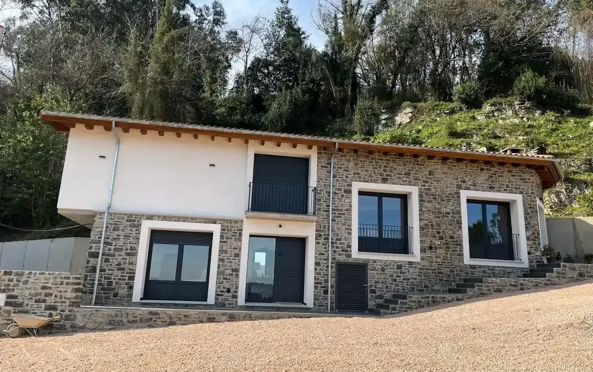 Casa de piedra y estuco blanco con ventanas y puertas negras en una ladera.