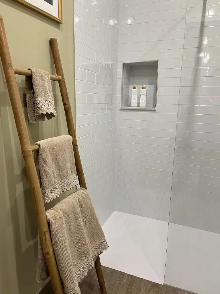 Baño con ducha de azulejos blancos, estante empotrado, toallas beige en una escalera de madera y pared verde oliva.