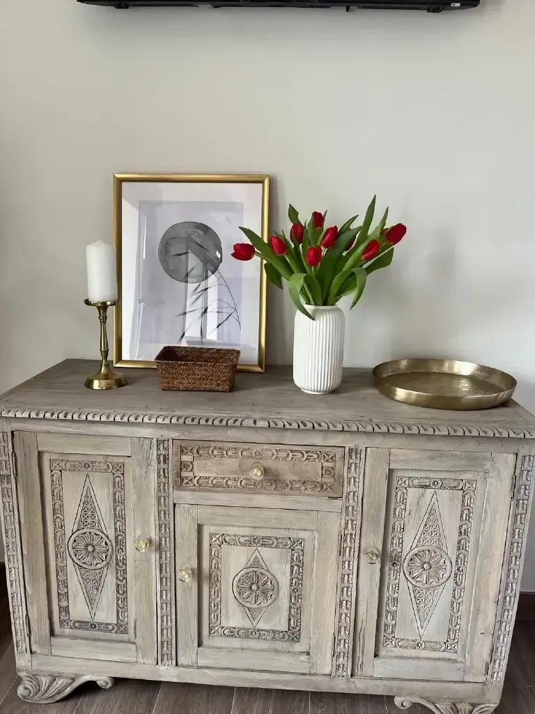 Mueble de madera tallada en color gris con decoración: obras de arte, flores, vela y bandeja dorada.