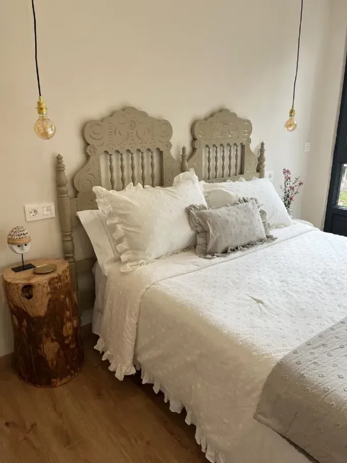 Dormitorio con cabecera gris ornamentada, ropa de cama blanca, mesa auxiliar con forma de tronco de árbol
