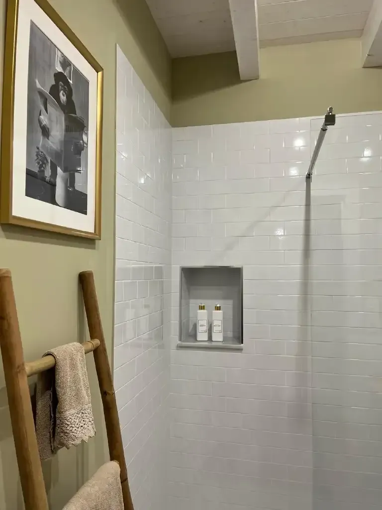 Baño con ducha de azulejos blancos, estante empotrado con botellas y escalera de madera con toallas.
