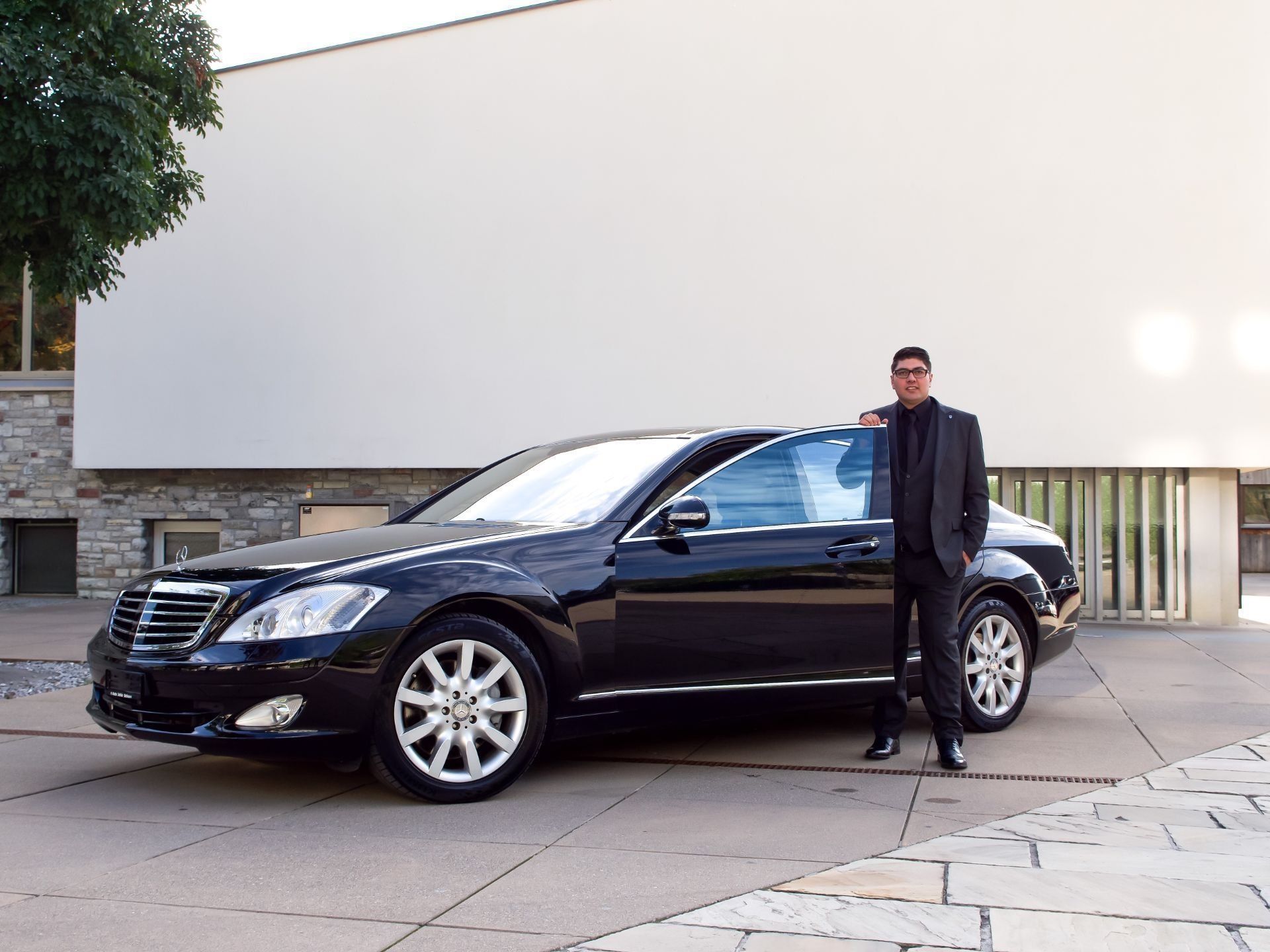 Ein Mann steht neben einer schwarzen Luxuslimousine, die vor einem modernen Gebäude geparkt ist.
