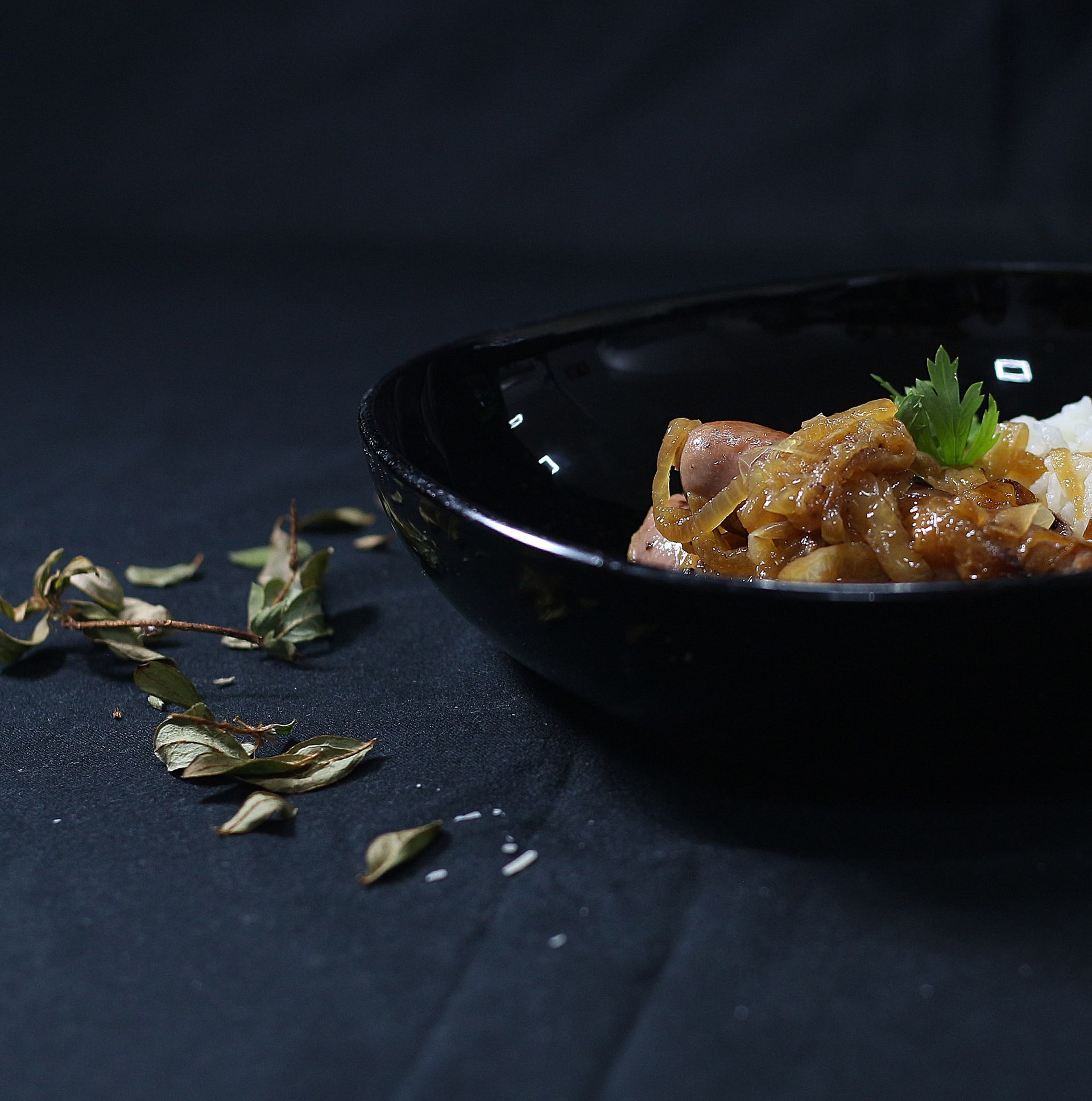 Cuenco negro de comida con cebollas, perejil y arroz sobre una superficie oscura con hierbas esparcidas.