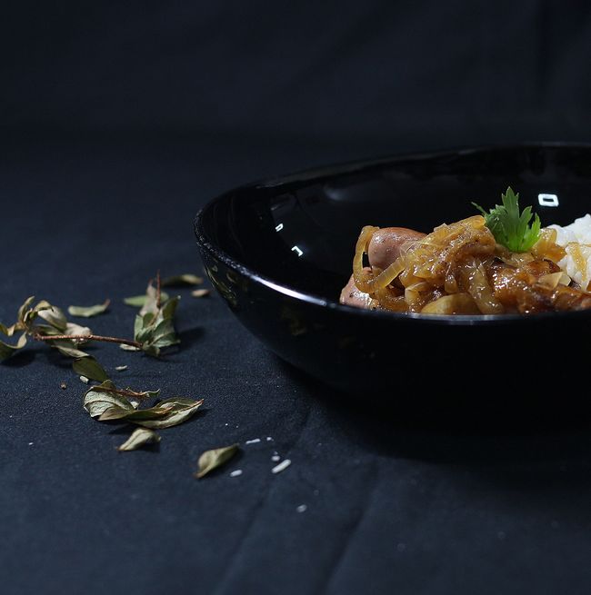 Cuenco negro de comida con cebollas, perejil y arroz sobre una superficie oscura con hierbas esparcidas.