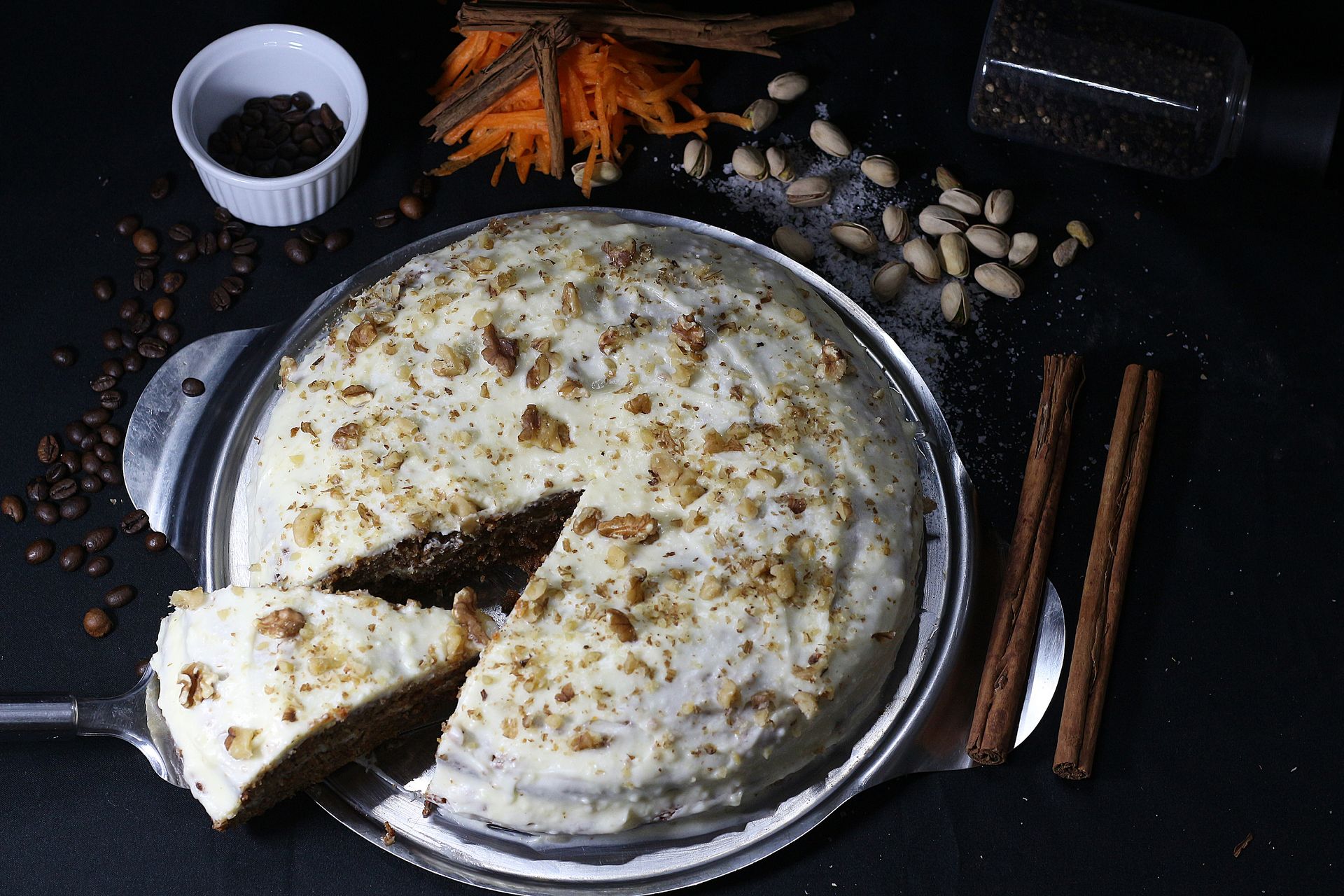 Pastel de zanahoria con glaseado de queso crema, adornado con nueces, granos de café y ramas de canela sobre una superficie negra.