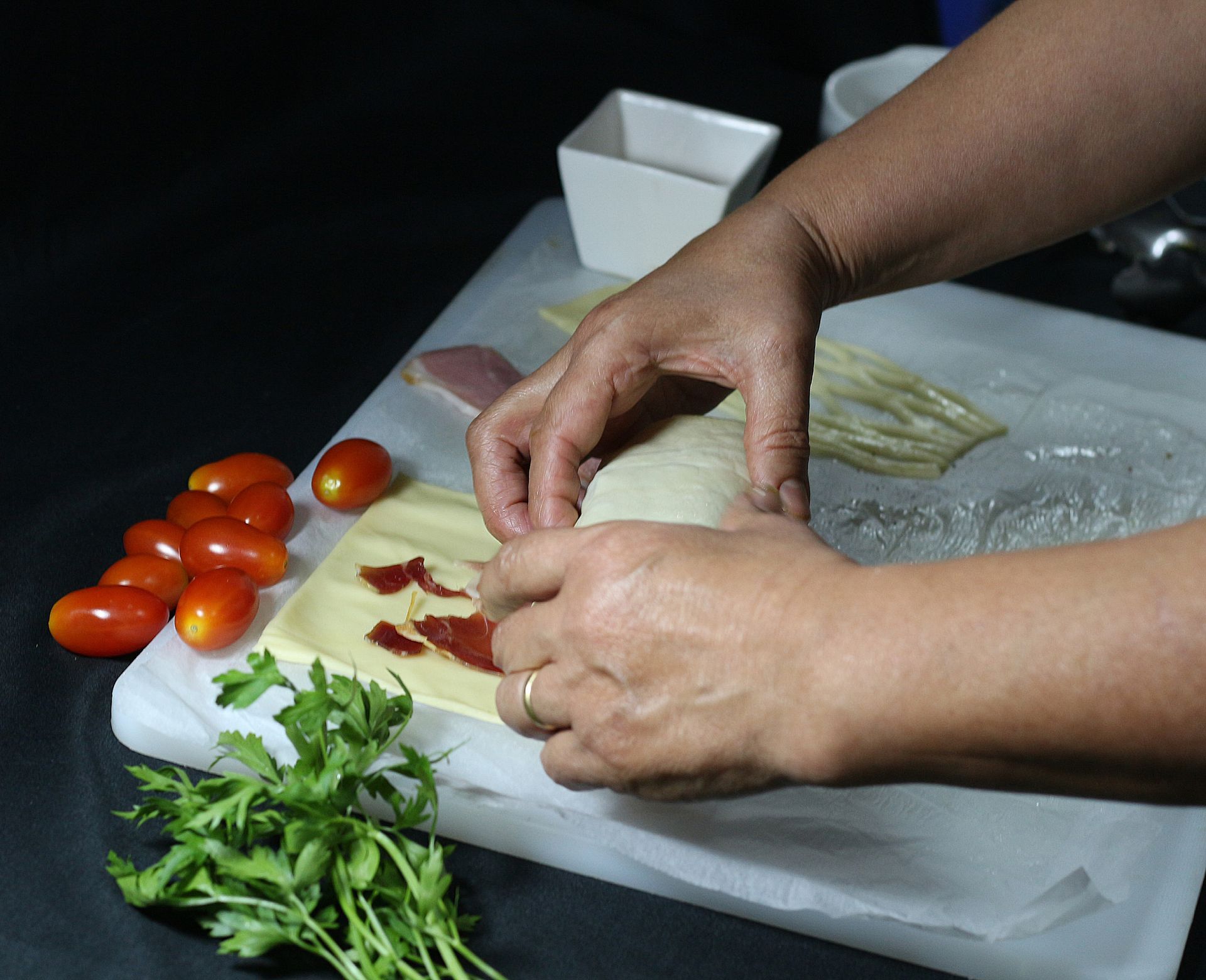 Manos haciendo rodar comida sobre papel pergamino con tomates y perejil cerca.