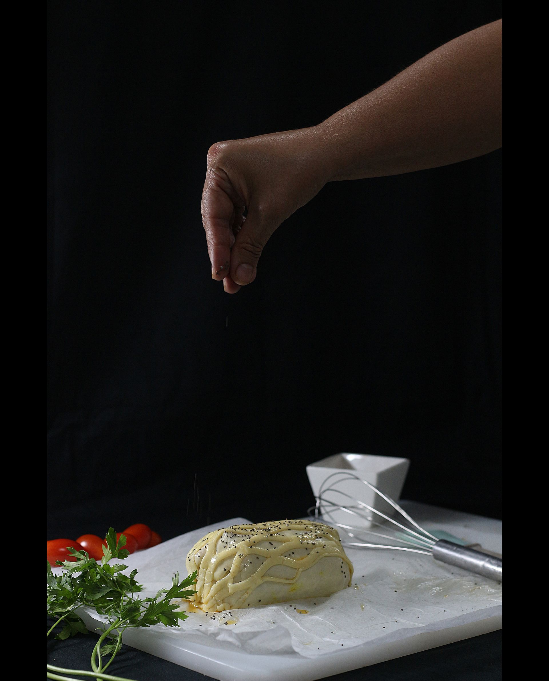 Espolvoreando a mano semillas de sésamo sobre la masa sobre una tabla de cortar, con un fondo negro.