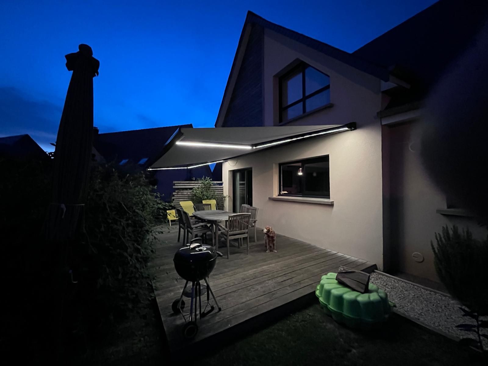 Patio à l’arrière de la maison au crépuscule, avec un auvent éclairé, un ensemble de salle à manger d’extérieur, un barbecue et la façade de la maison.