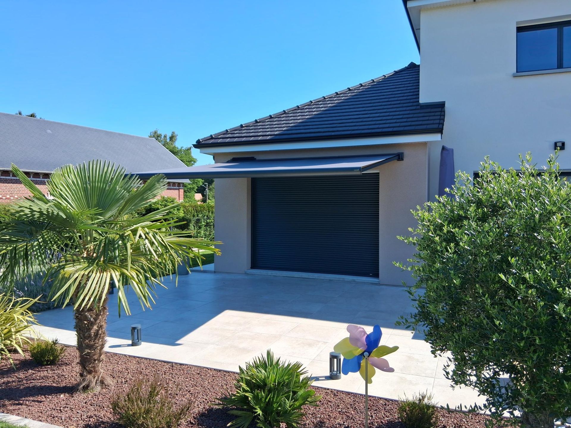 Extérieur d’une maison moderne avec un toit sombre, une porte de garage, un palmier et un petit jardin devant la maison