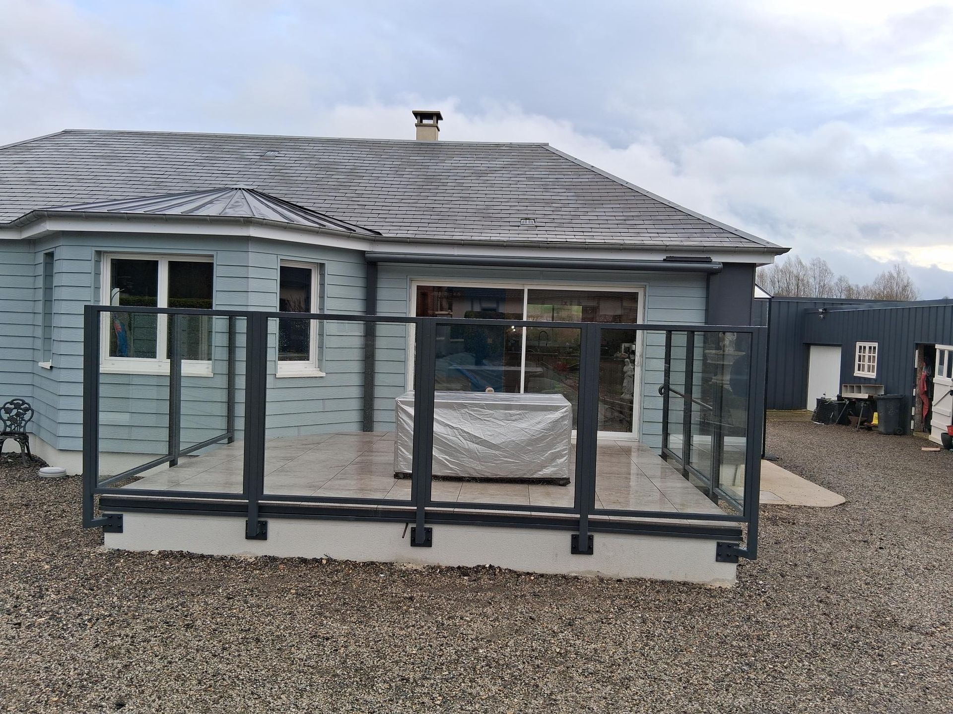 Petite maison bleue avec une terrasse vitrée sur un terrain gravillonné