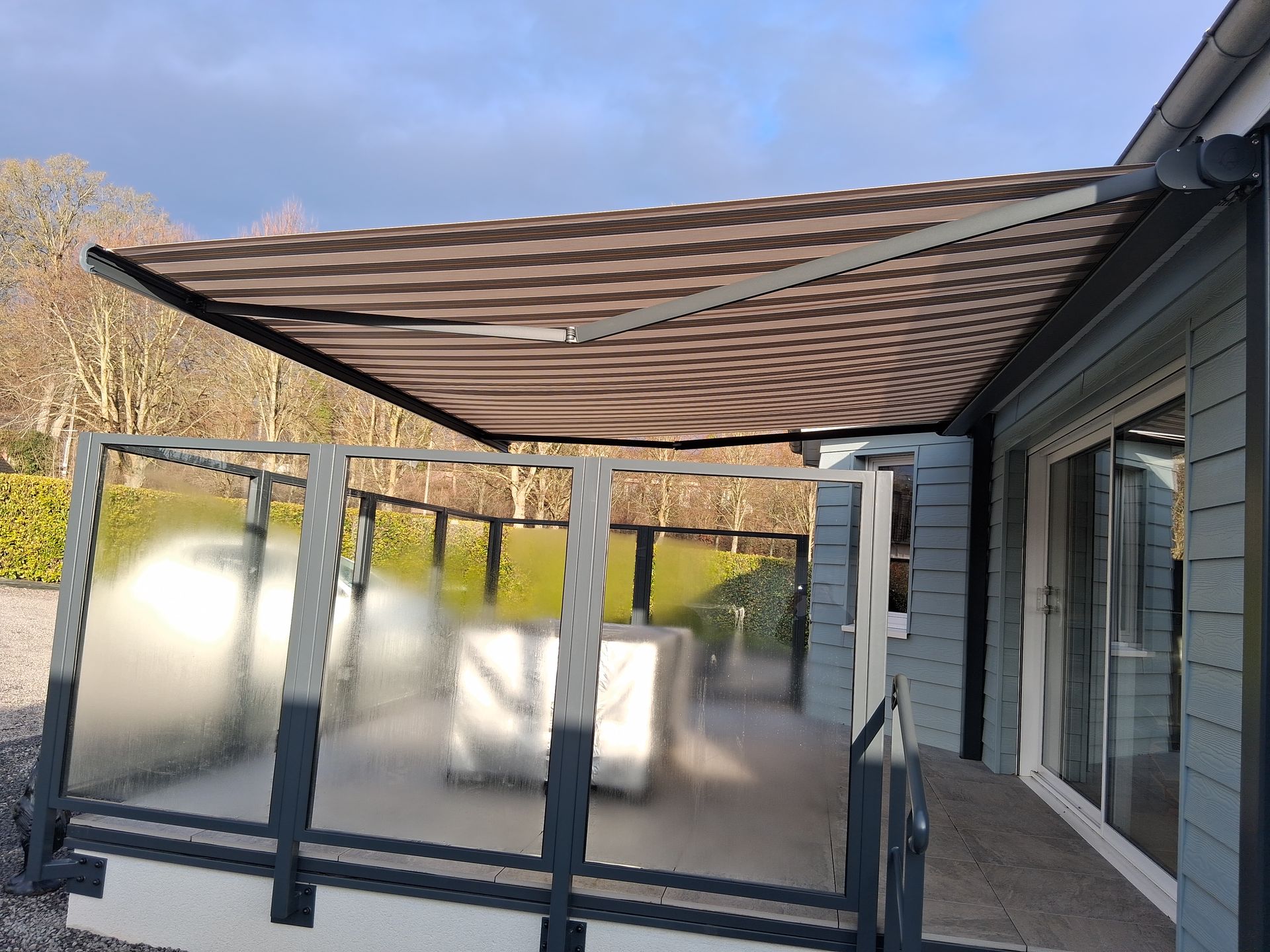 Patio couvert avec un store rayé et une rambarde en verre dépoli, à côté d’une maison.