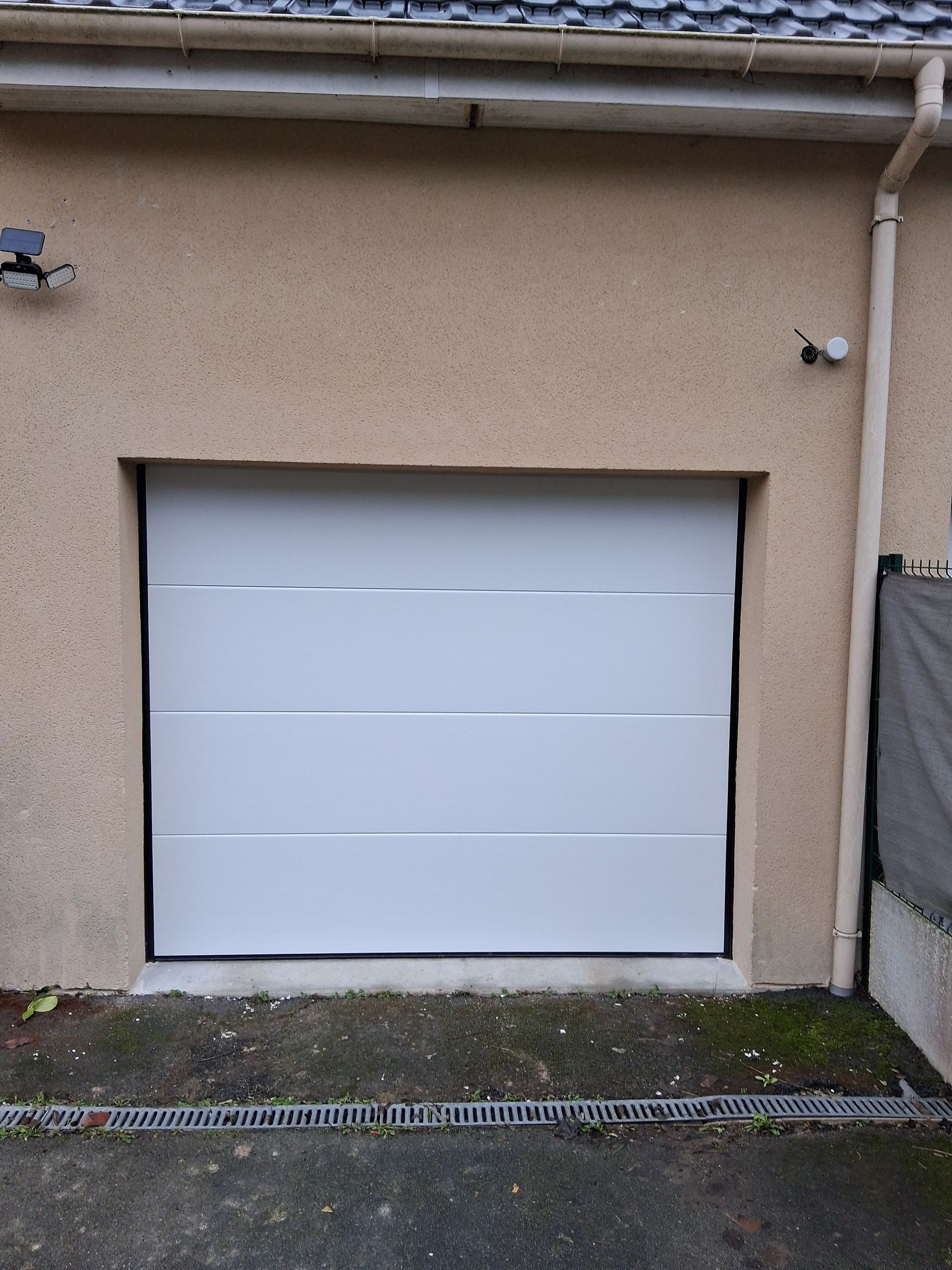 Porte de garage blanche encastrée dans un mur extérieur beige, avec une petite caméra de surveillance au-dessus.