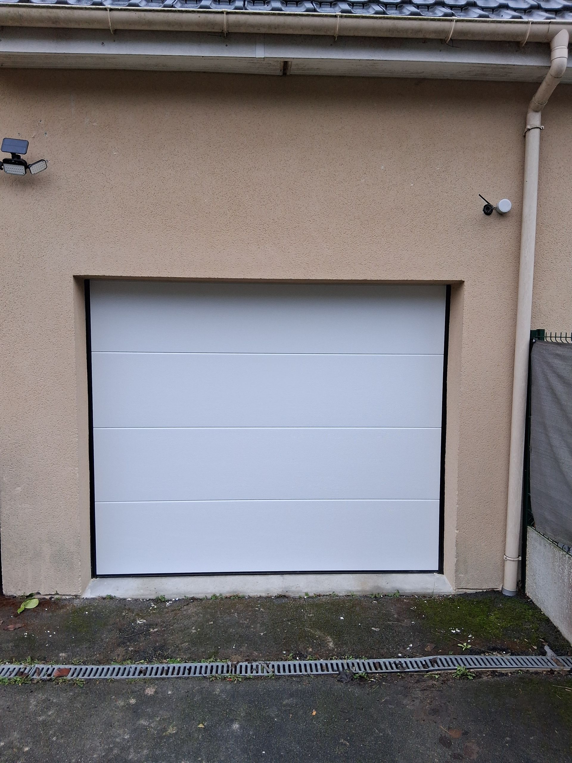 Porte de garage blanche intégrée dans un mur en stuc beige, avec une petite lampe au-dessus et du gravier au pied du mur.