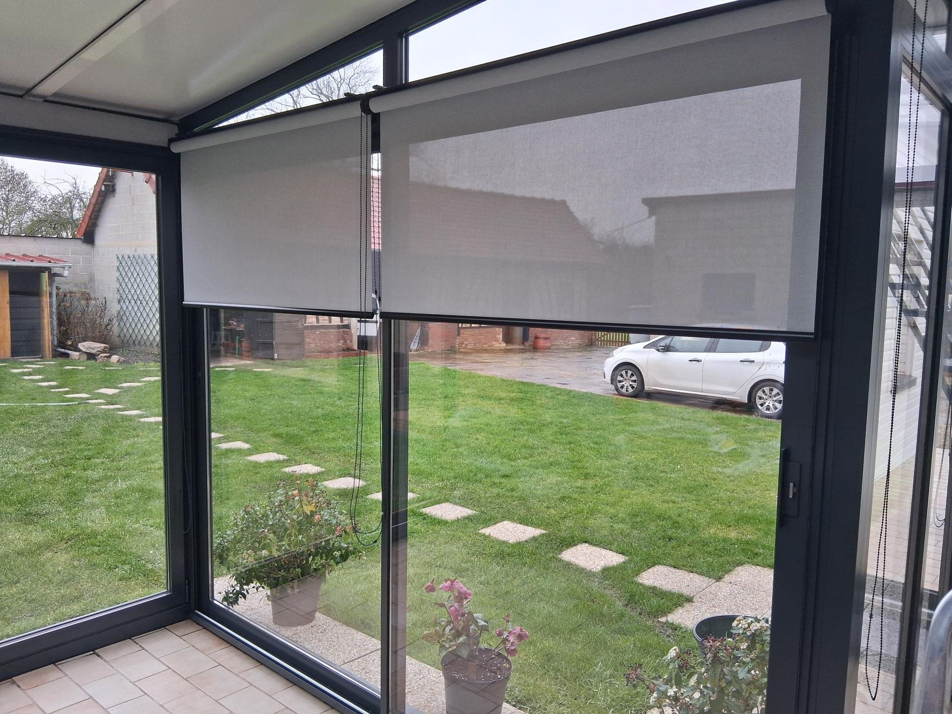 Patio fermé, avec des stores enrouleurs gris, des parois vitrées et une vue sur une pelouse et une voiture blanche garée.
