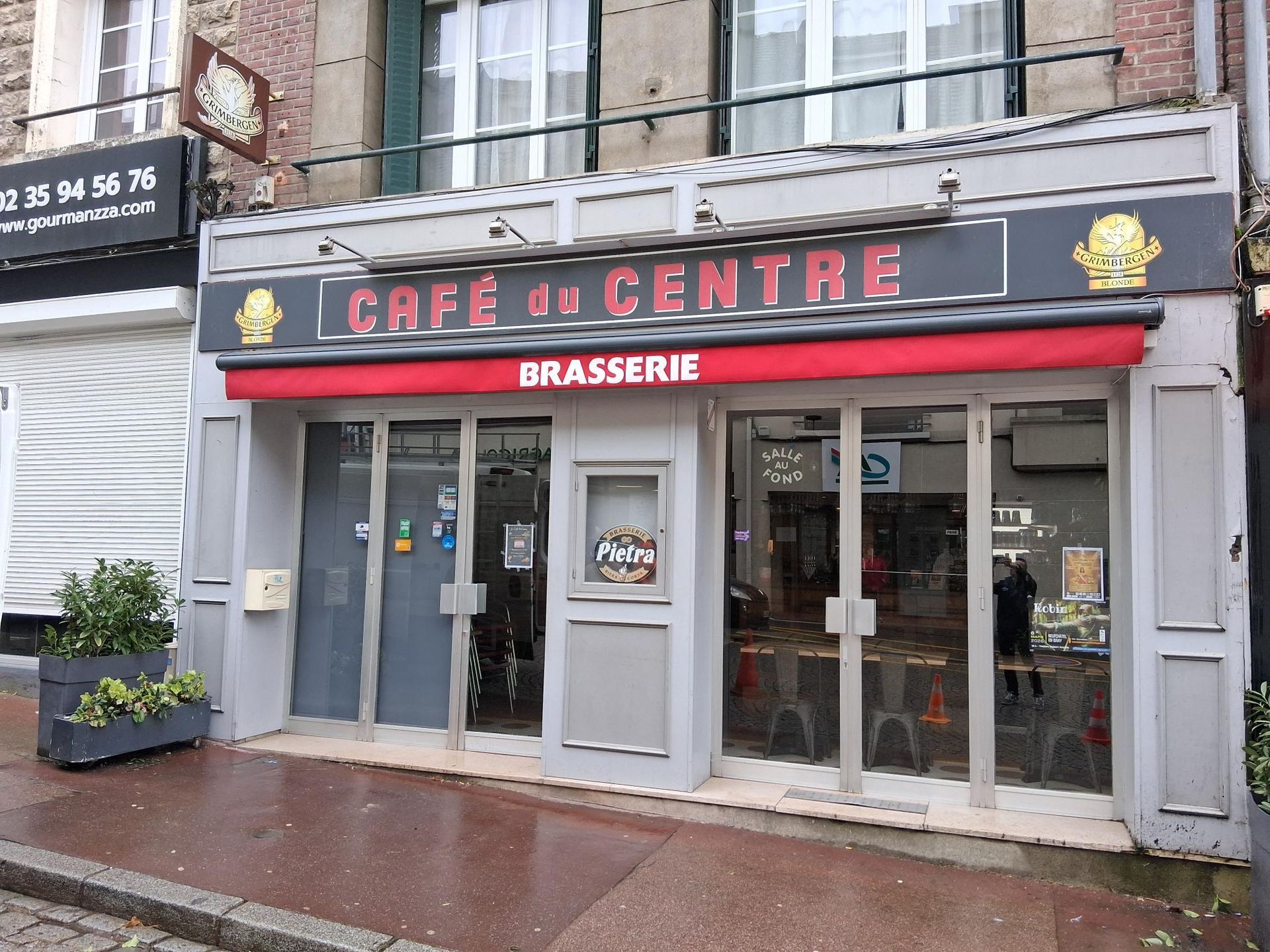 Vue d’une rue pluvieuse avec la brasserie Café du Centre, sa devanture grise et son auvent rouge.