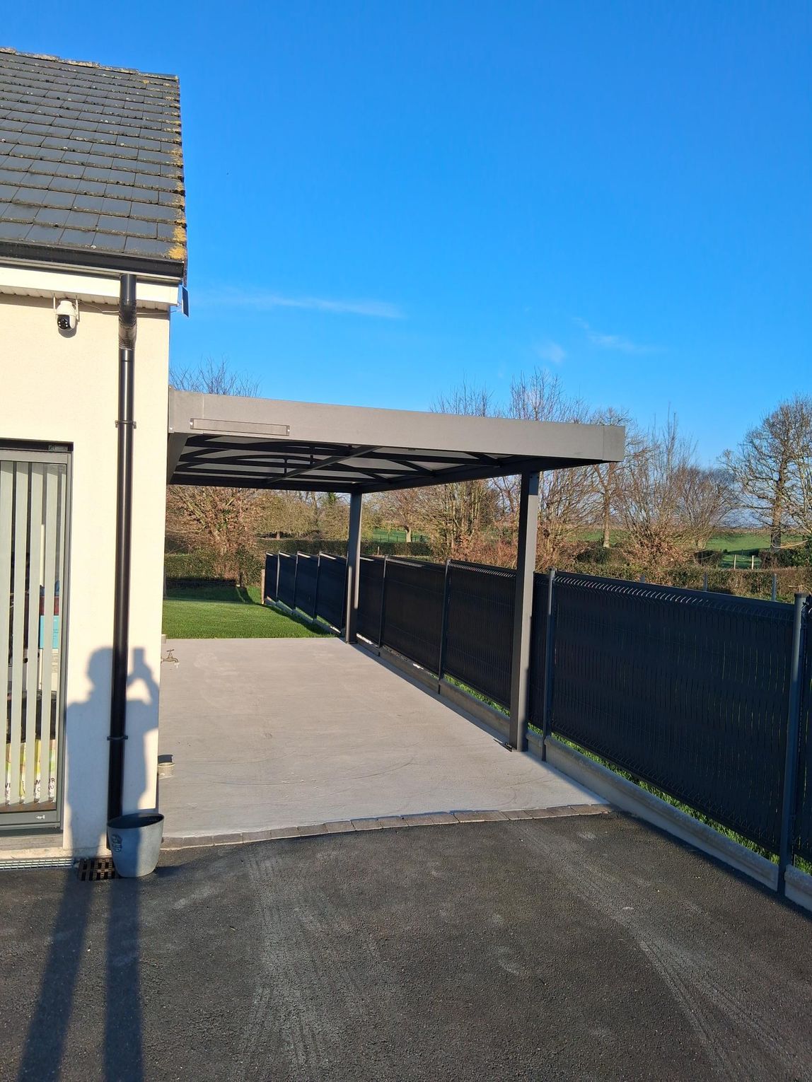 Allée couverte à côté d’une maison, avec une clôture en métal noir et un ciel bleu dégagé