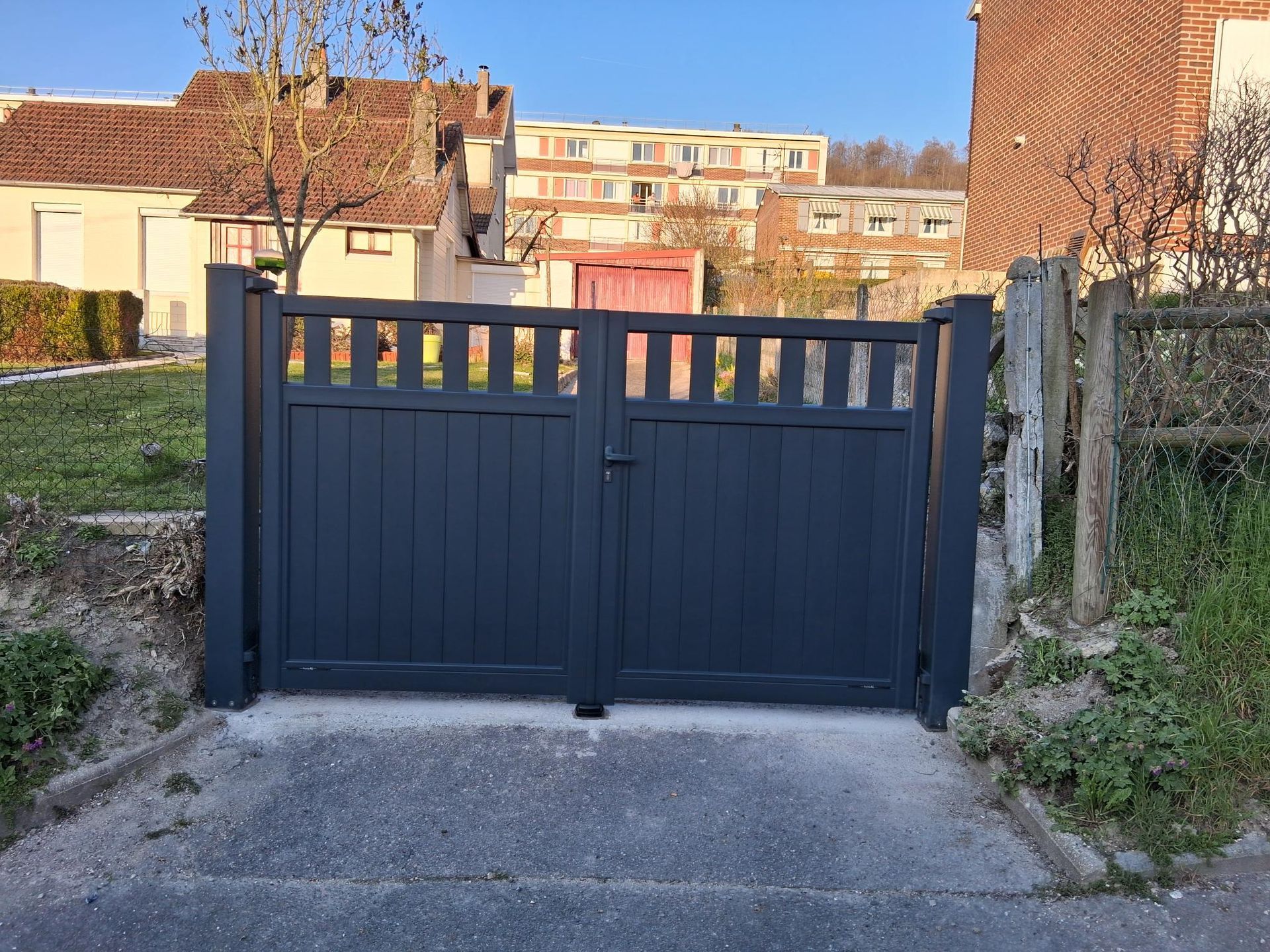 Portail métallique bleu foncé bloquant une allée étroite entre des bâtiments et une végétation envahissante