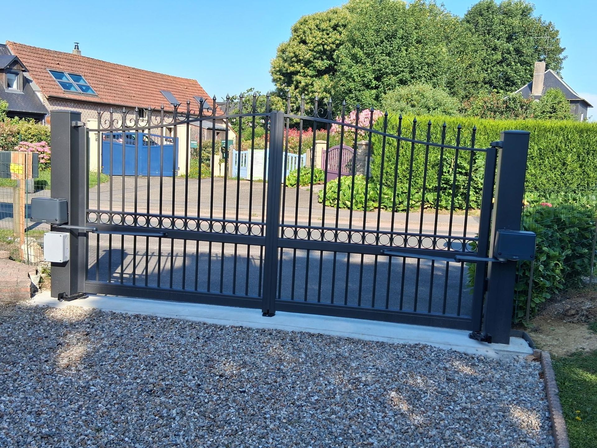 Portail d’allée en métal noir avec des piliers en pierre, à côté d’un chemin de gravier, et une maison en arrière-plan