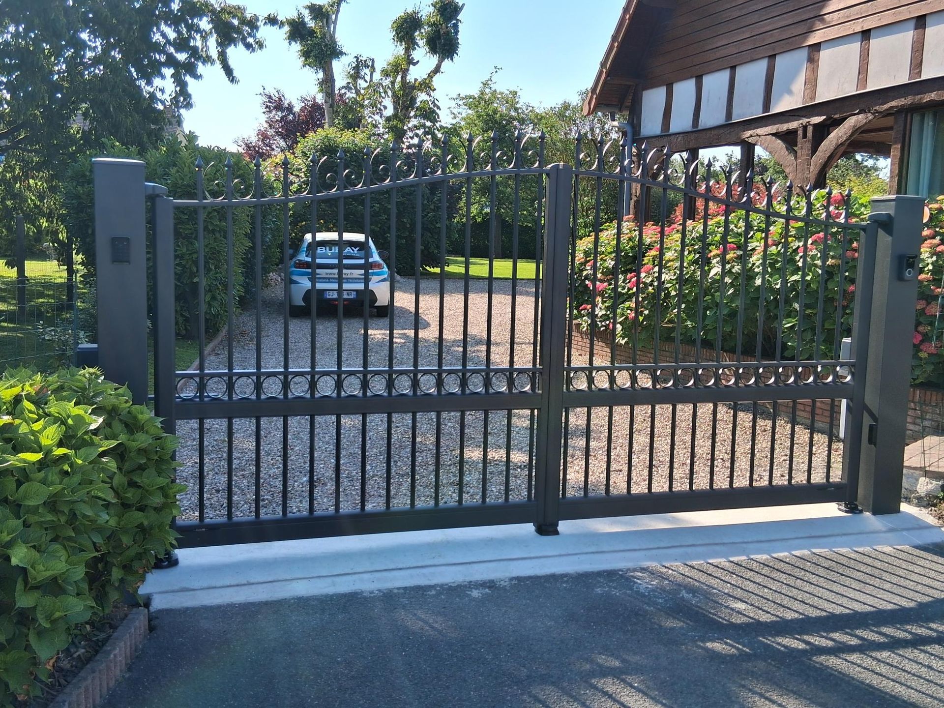 Portail d’allée en métal noir, avec une voiture bleue derrière et une terrasse couverte sur la droite