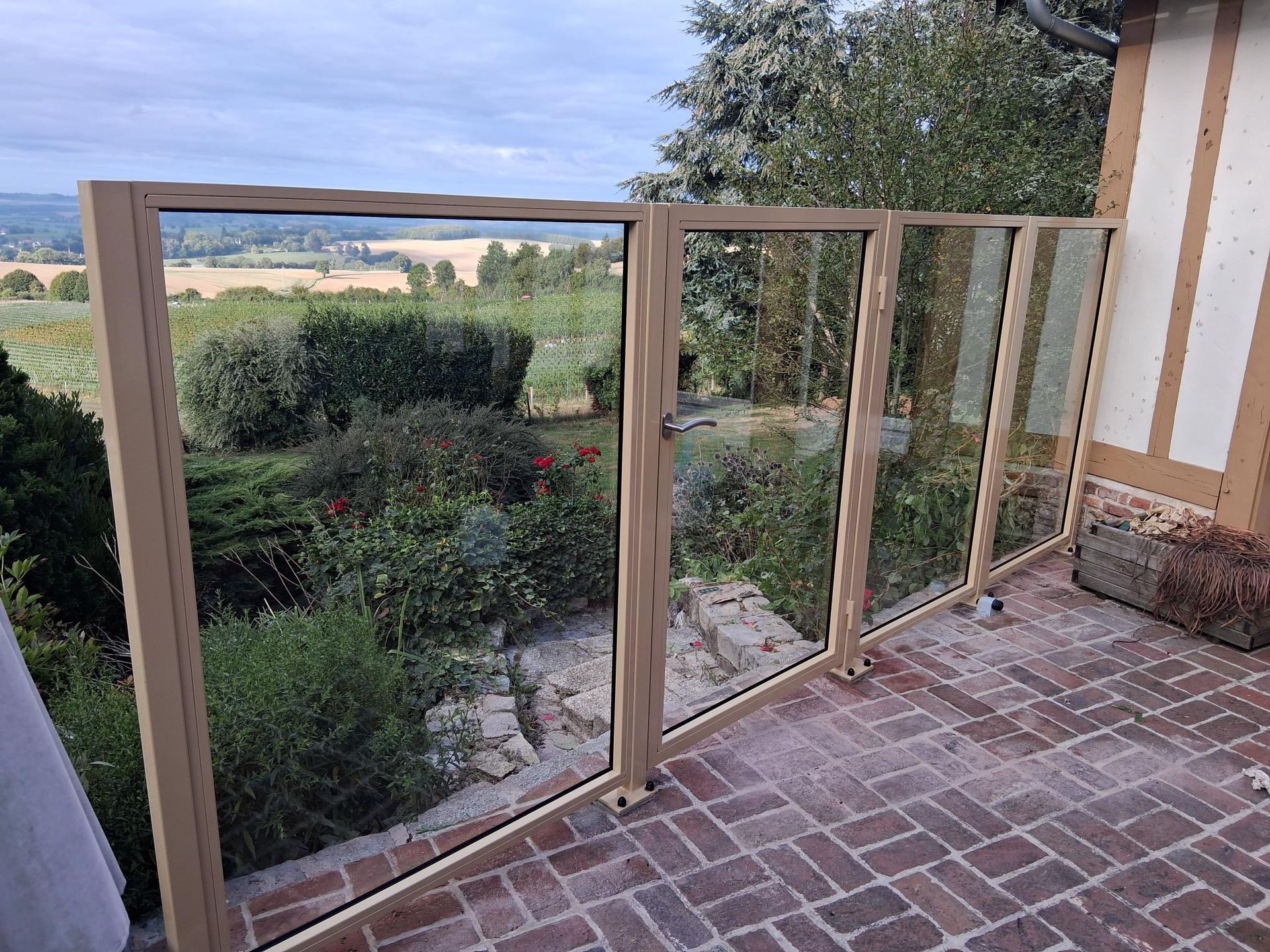 Brise-vent en verre encadré de bois, le long d’une terrasse en briques donnant sur un jardin et des champs à perte de vue