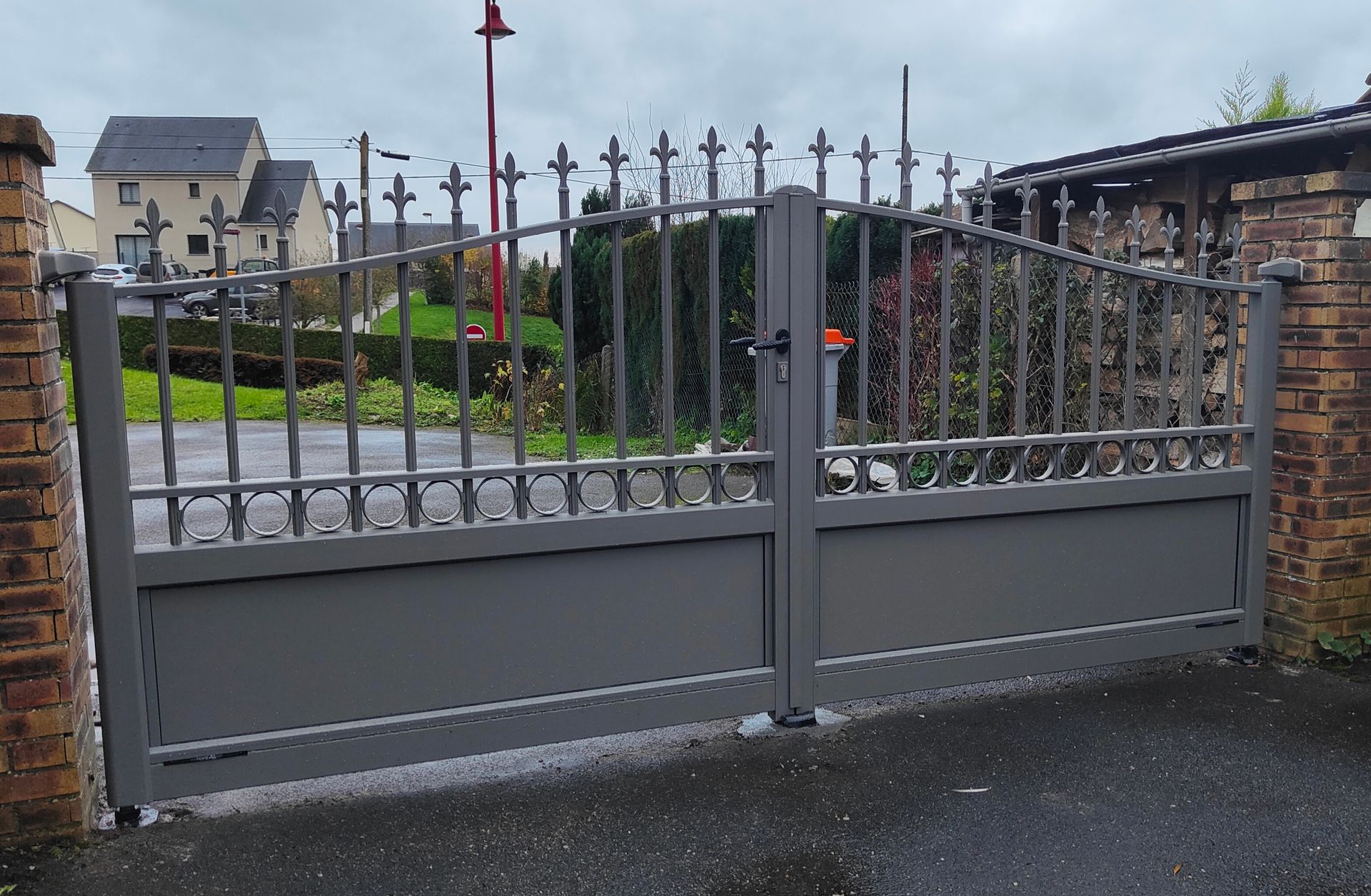 Portail d’allée gris en métal à barreaux pointus, encadré de murs en briques, dans un quartier résidentiel
