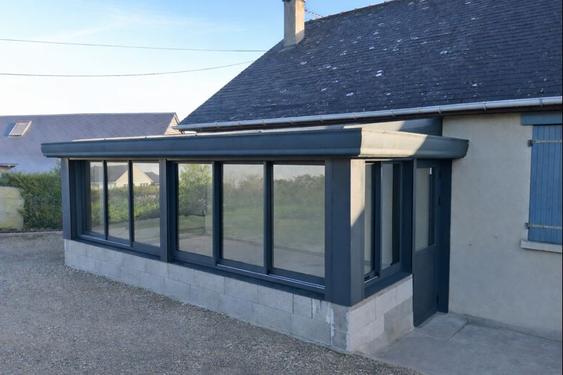 Extension de maison contemporaine avec véranda vitrée et cadre sombre, donnant sur une allée en gravier