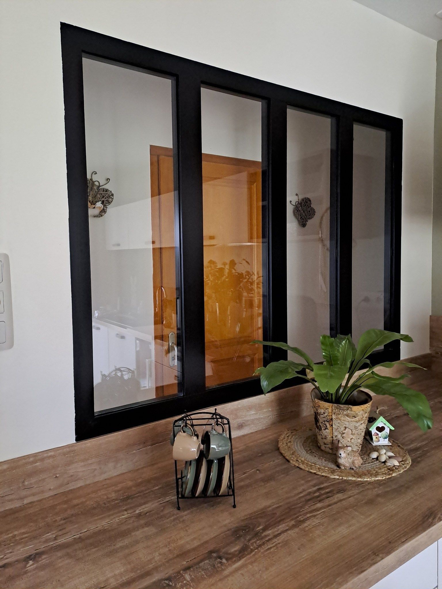 Fenêtre intérieure à cadre noir au-dessus d’un comptoir en bois, avec une plante en pot et un bougeoir.