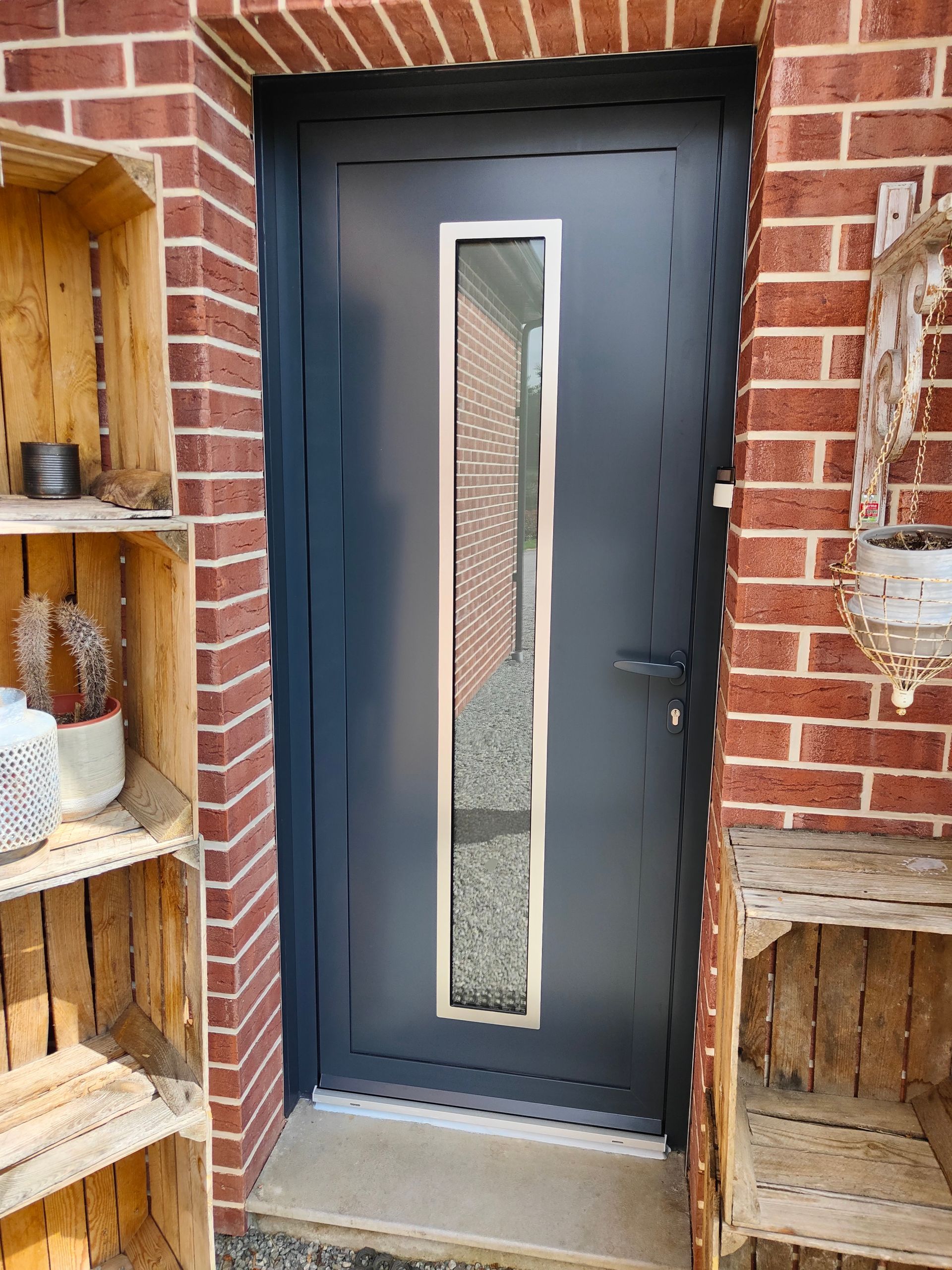 Porte gris foncé avec un étroit panneau vitré dépoli dans une entrée en briques rouges.