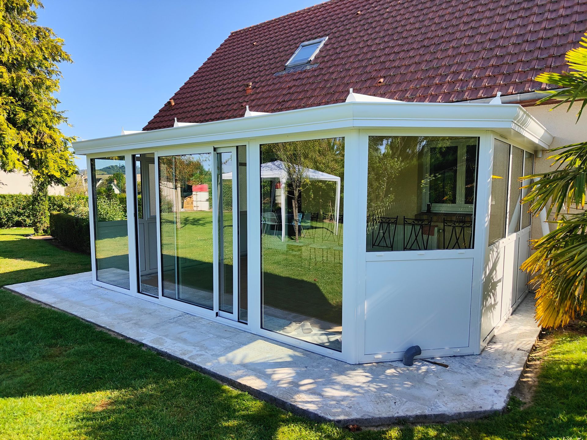 Véranda en verre blanc attenante à une maison, avec des portes coulissantes et des fenêtres donnant sur une pelouse verdoyante.
