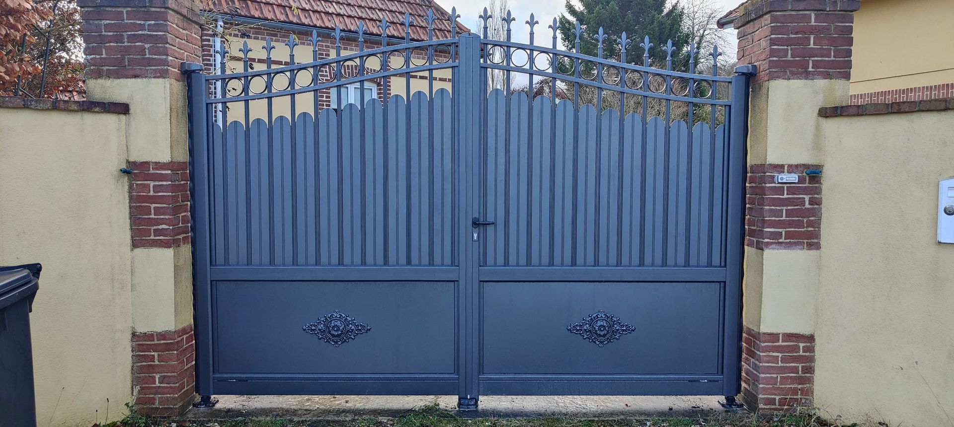 Portail métallique bleu à deux battants devant une maison en briques et des murs couleur crème