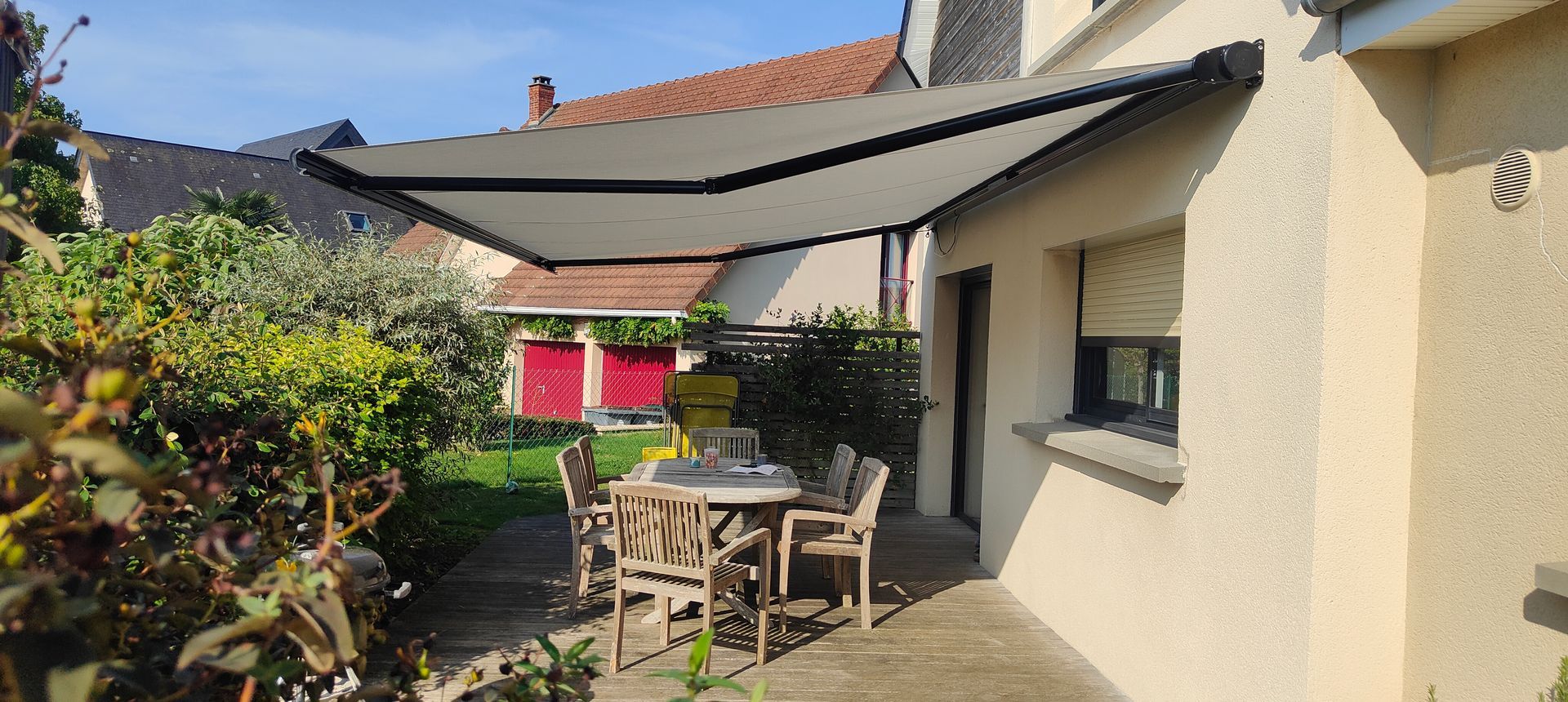 Patio avec une table et des chaises sous un auvent rétractable, à côté d’une maison beige et de plantes de jardin
